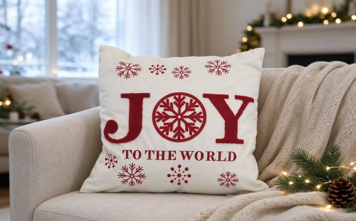 A square white throw pillow with red embroidered text reading 'JOY TO THE WORLD' and red snowflakes, resting on a beige couch in a decorated living room.