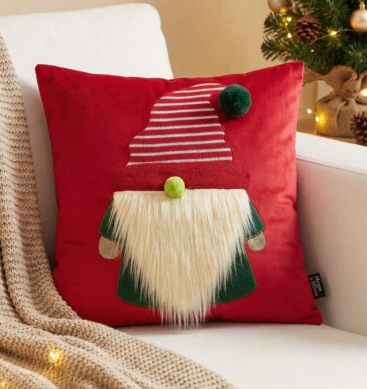 A red square throw pillow featuring a gnome applique with a white faux-fur beard, green clothing, and a striped red-and-white hat, styled on a white armchair with a beige knit blanket and a softly blurred Christmas tree in the background.