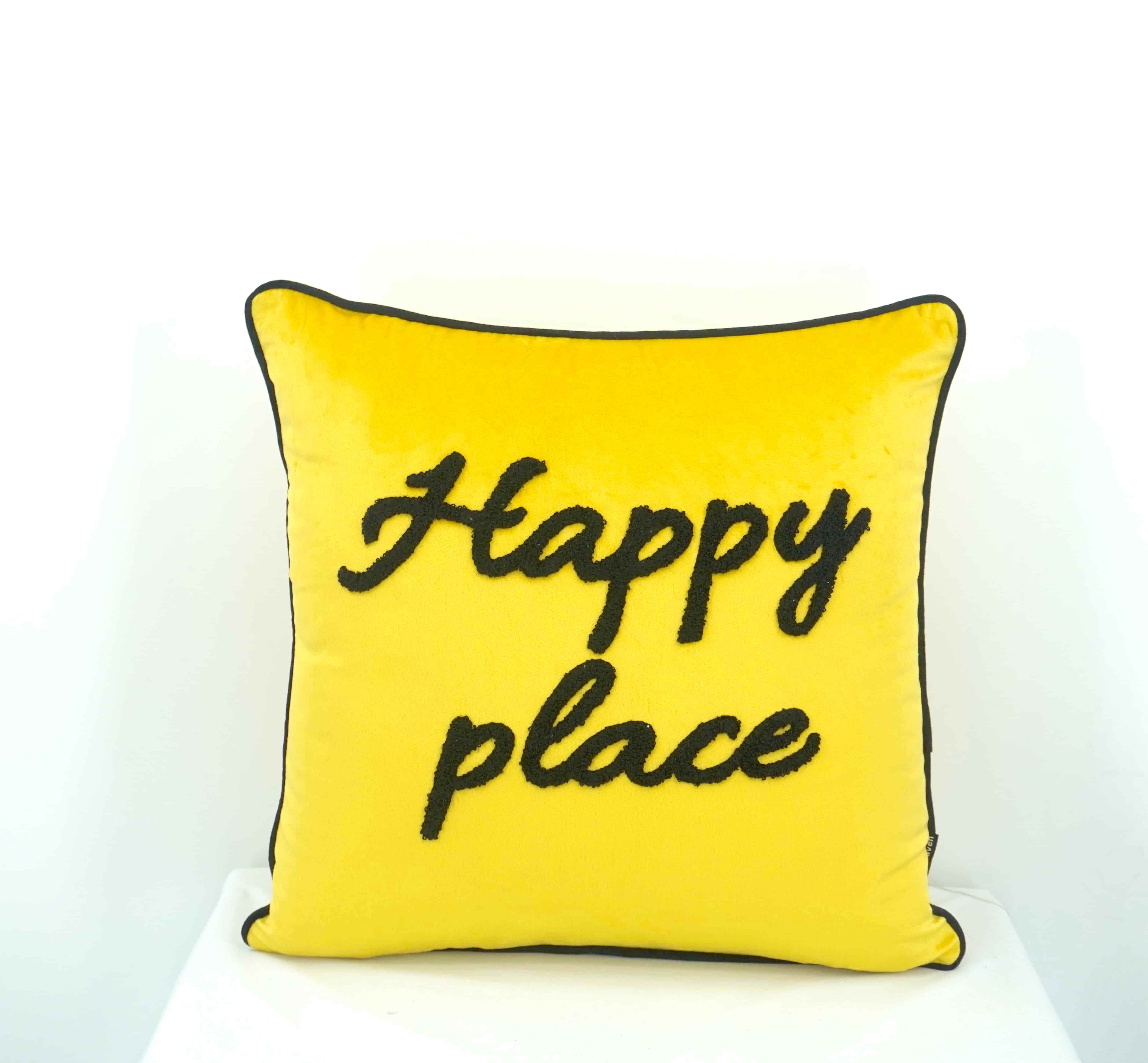 A square yellow throw pillow with black piping and the words 'Happy place' embroidered in black script on the front, displayed against a clean white background.