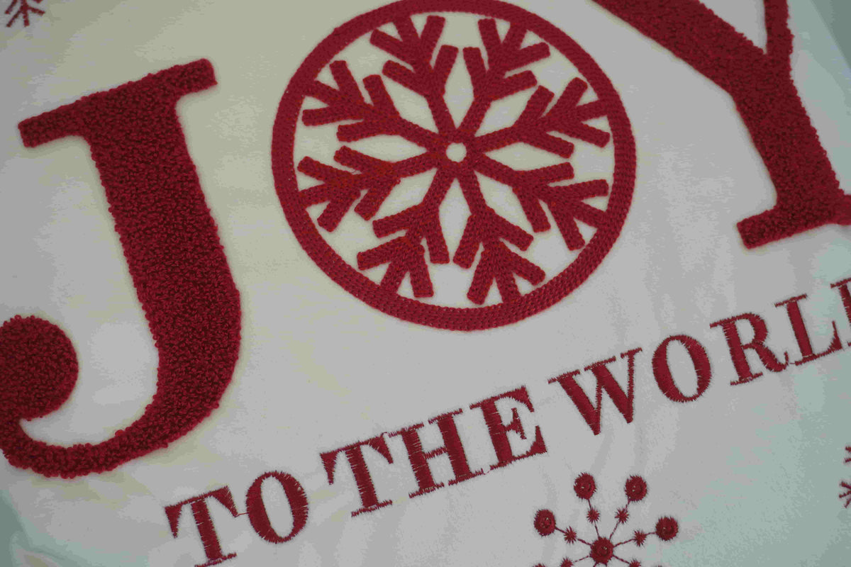 A close-up of a festive textile or printed design featuring the word "JOY" in large red letters with a red snowflake inside the O, and the phrase "TO THE WORLD" below on a white background.