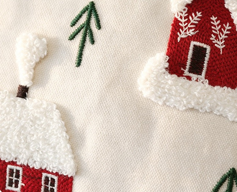 Close-up of a textured cream fabric featuring embroidered winter cottages in red and white with dark green pine trees.