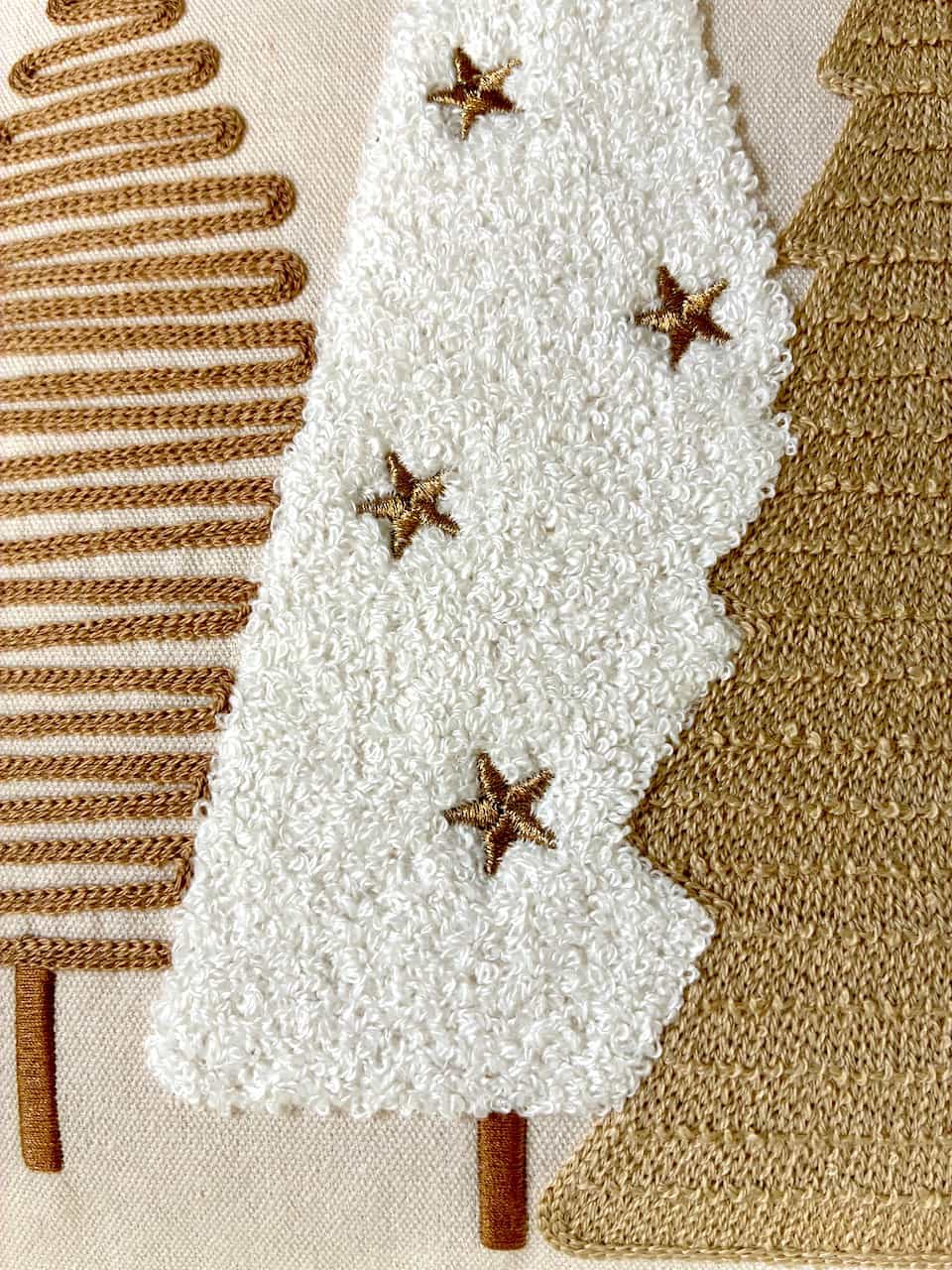 Close-up of a textured fabric artwork featuring stylized Christmas trees in white, tan, and brown, with embroidered star accents.