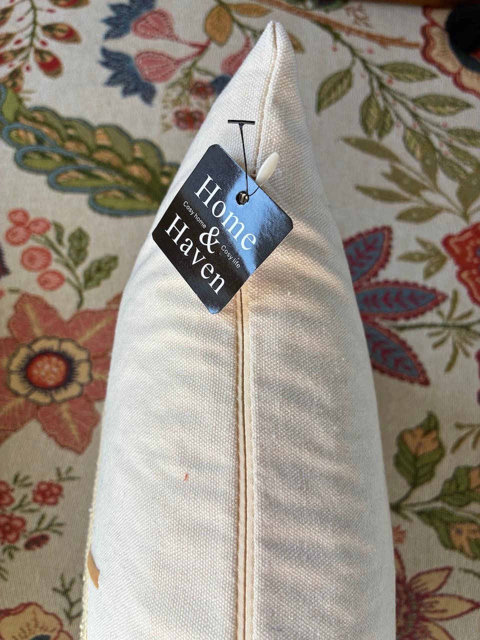 An off-white woven throw pillow with a black 'Home & Haven' brand tag, resting on a floral-patterned fabric surface.