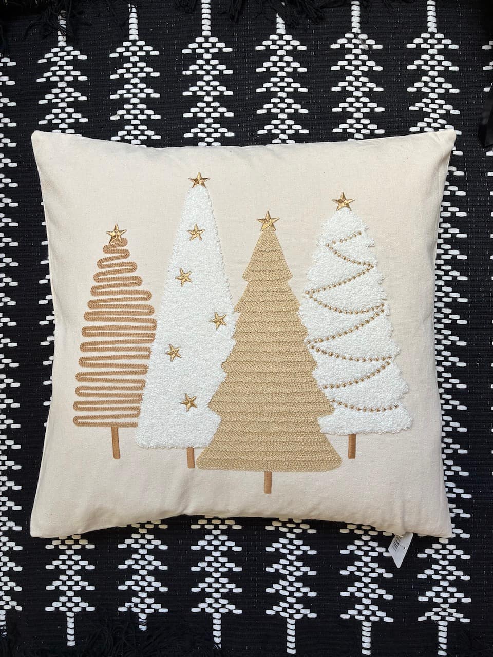 A square cream throw pillow featuring three stylized Christmas tree appliqués in tan and white with small gold stars, displayed on a black and white patterned woven rug.