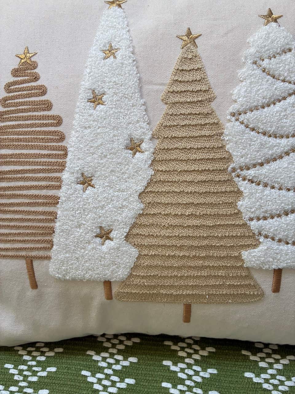A close-up of a decorative throw pillow featuring embroidered Christmas tree shapes in beige and white with gold star accents, resting on a green and white patterned rug.
