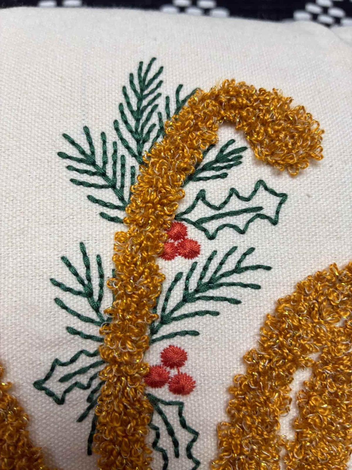 Close-up of a white woven fabric featuring a gold glittery embroidered candy cane with green holly leaves and small red berries.