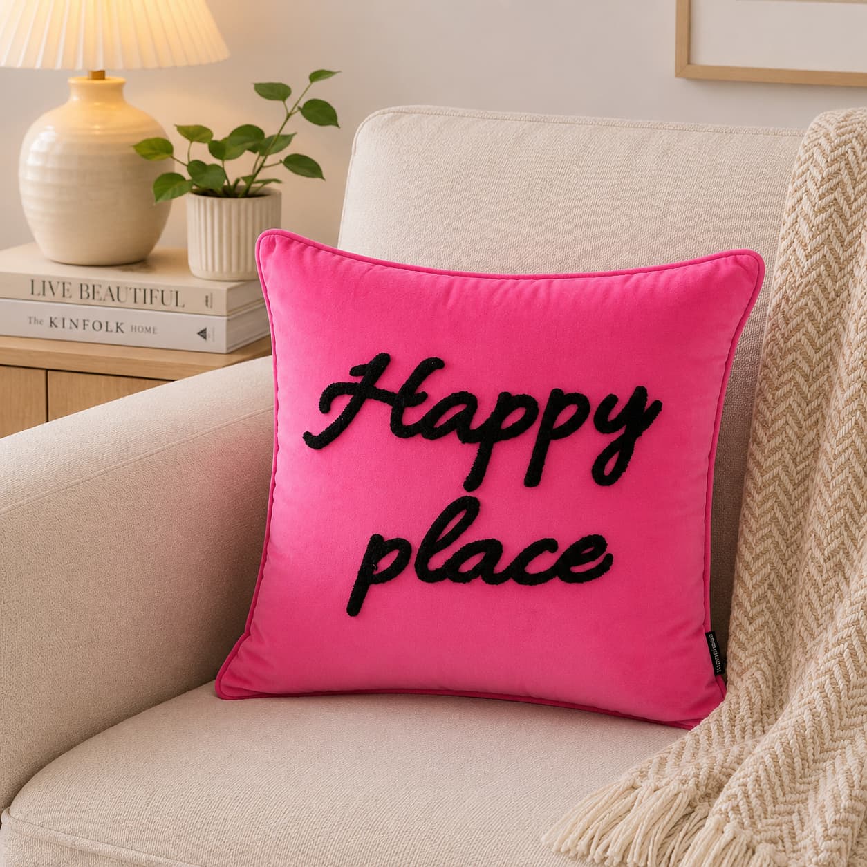 A bright pink square throw pillow with the words 'Happy place' embroidered in black script, resting on a beige armchair in a cozy living room setting.