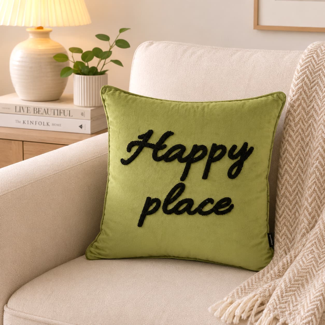 An olive green throw pillow with the words 'Happy place' embroidered in black script, resting on a cream-colored armchair in a cozy living room setting.