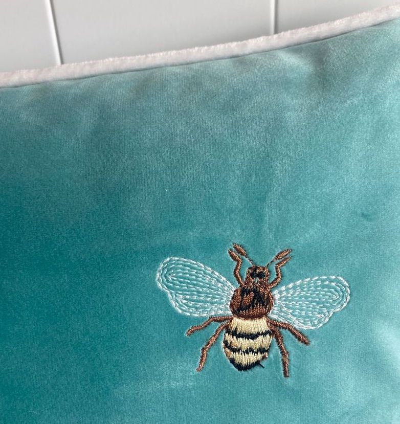 Close-up detail of a realistic embroidered bee with translucent white thread wings and yellow striped body, stitched on a soft teal velvet cushion cover.