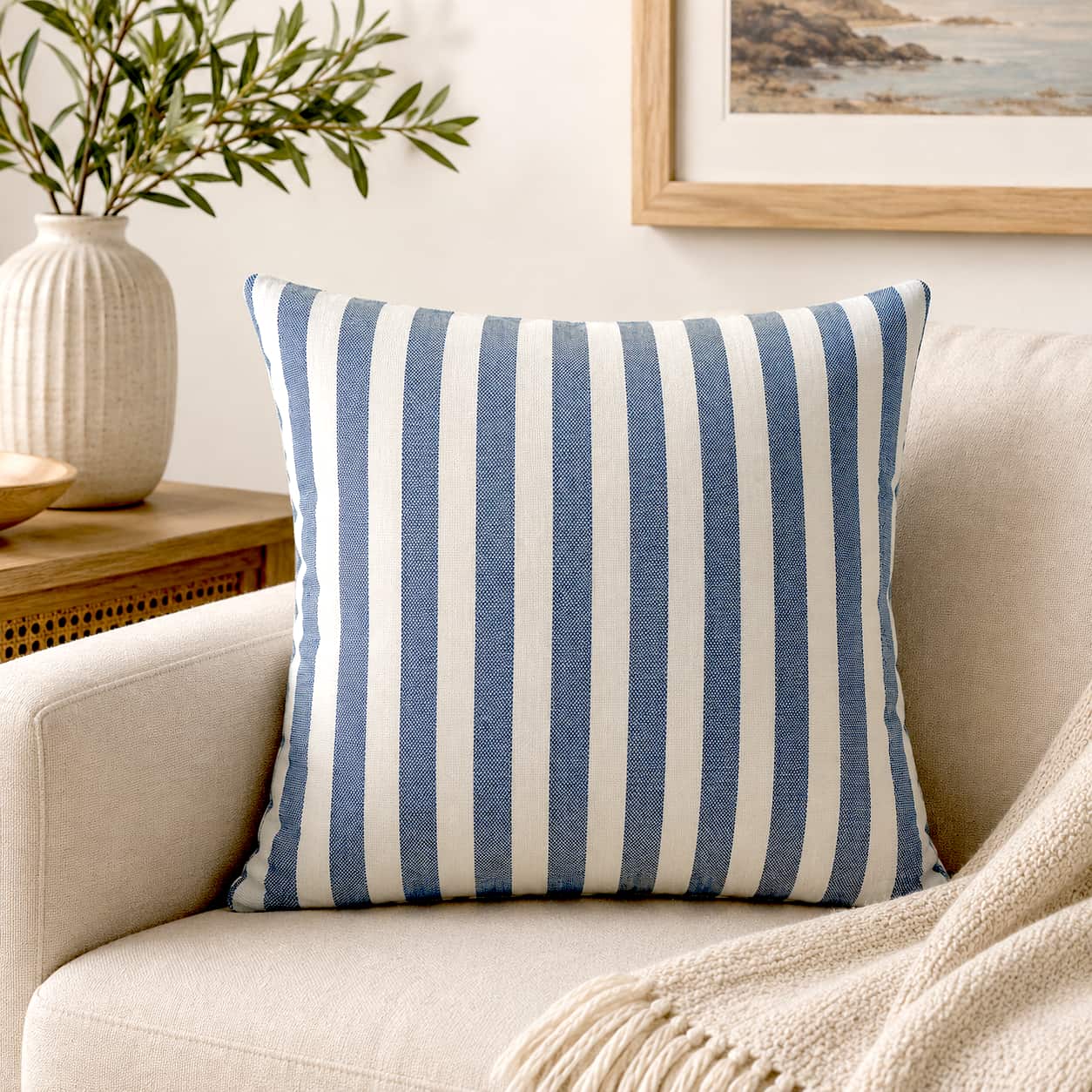 Blue and cream vertical striped throw pillow with a coastal style, placed on a beige sofa with a fringe blanket and a vase of green branches nearby.