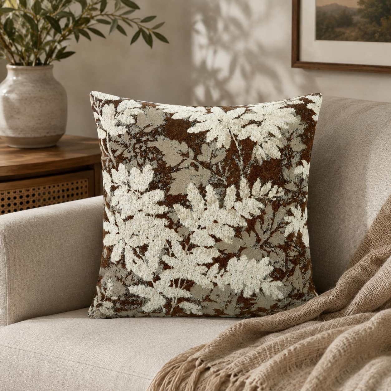 Rich brown and cream woven leaf pattern throw pillow, styled on a beige sofa with a fringe blanket and a vase of green branches.