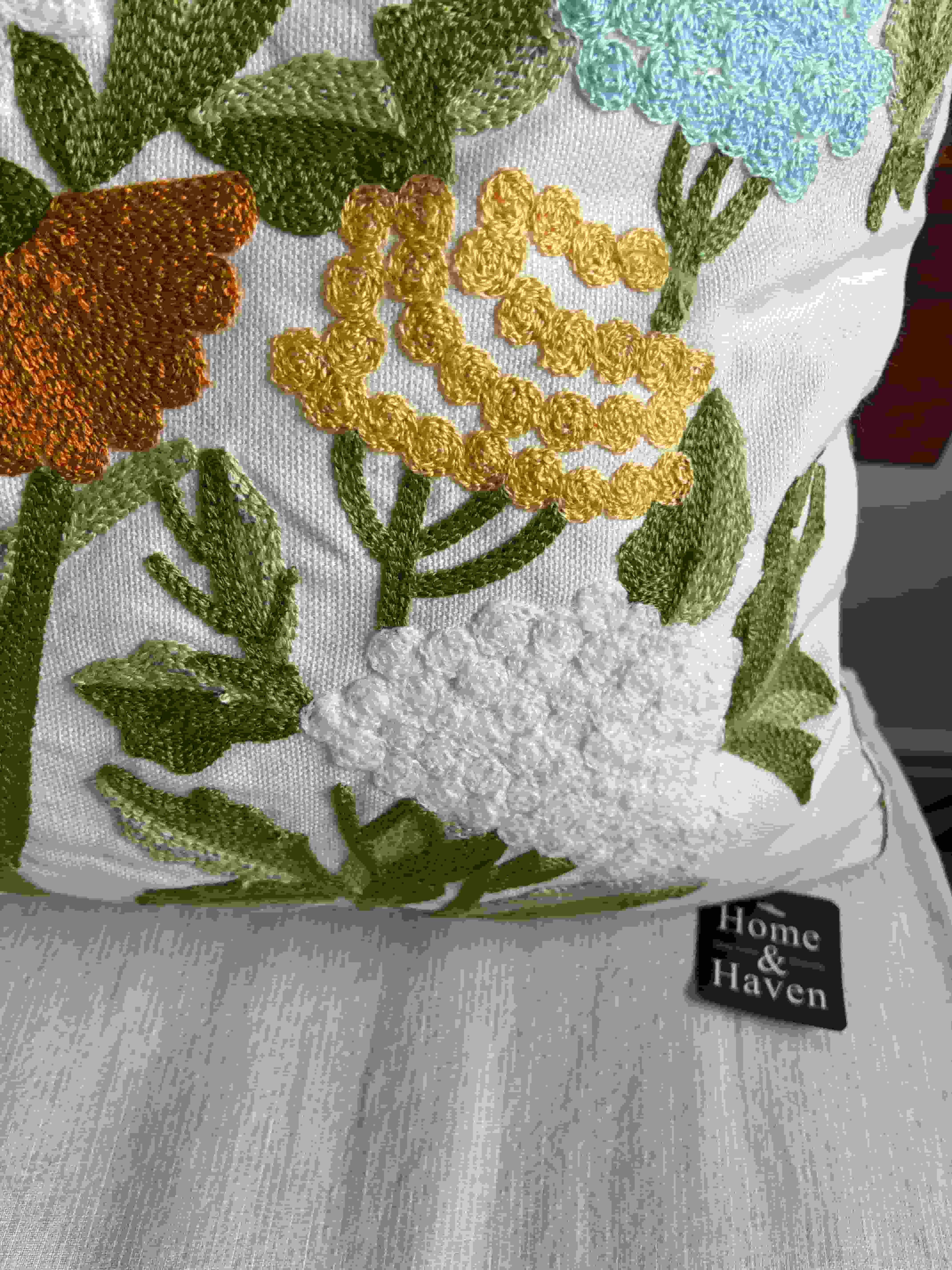 Macro shot of yellow, white, and blue textured embroidered flowers next to the black Home & Haven logo label on a white throw pillow.