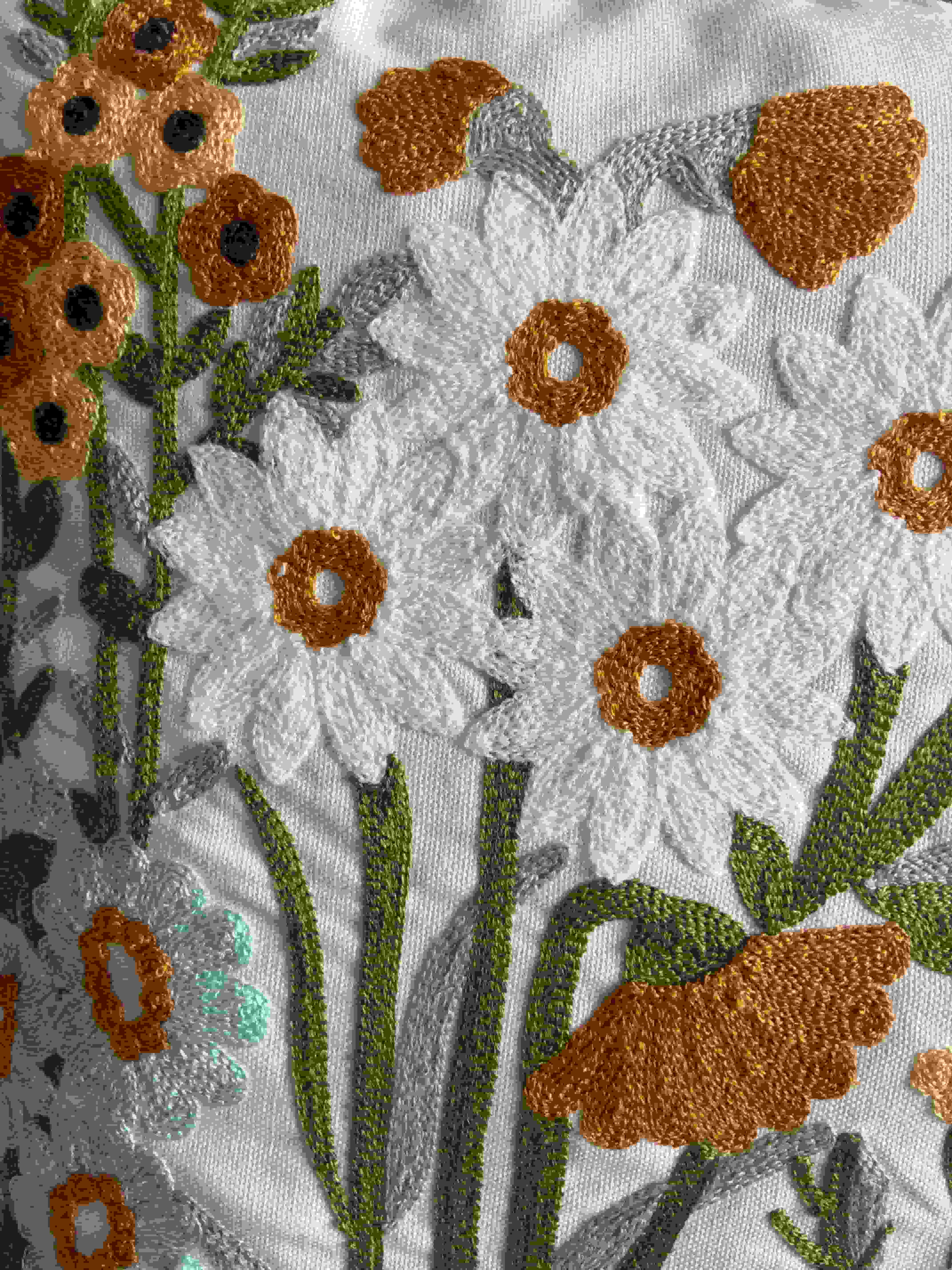Close-up detail of white daisy flowers and orange blooms with textured, raised embroidery stitching on a cream fabric cushion cover.


