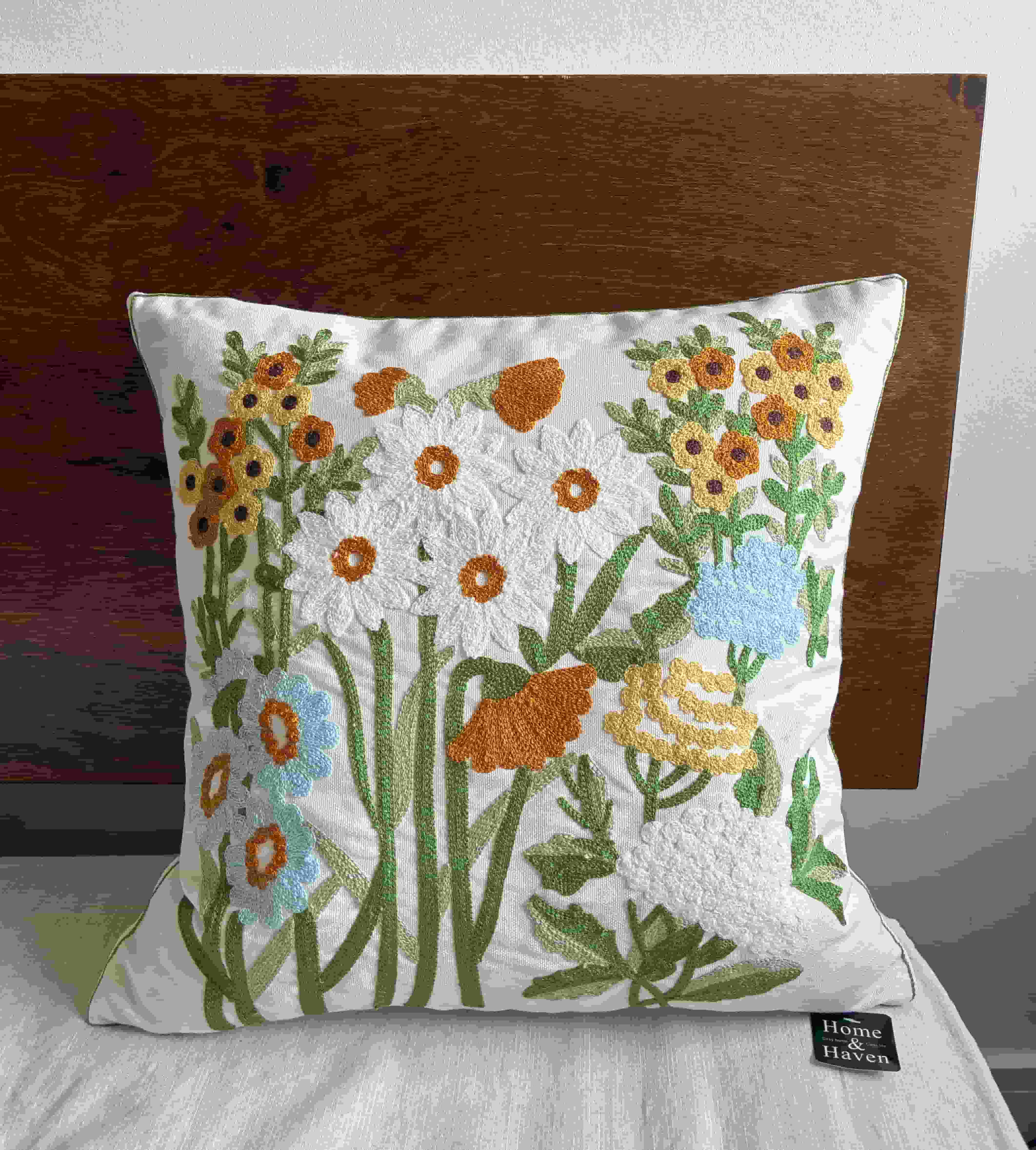 Ivory botanical embroidered cushion cover standing upright on a white bed with a dark brown wooden headboard, with a Home & Haven logo tag.
