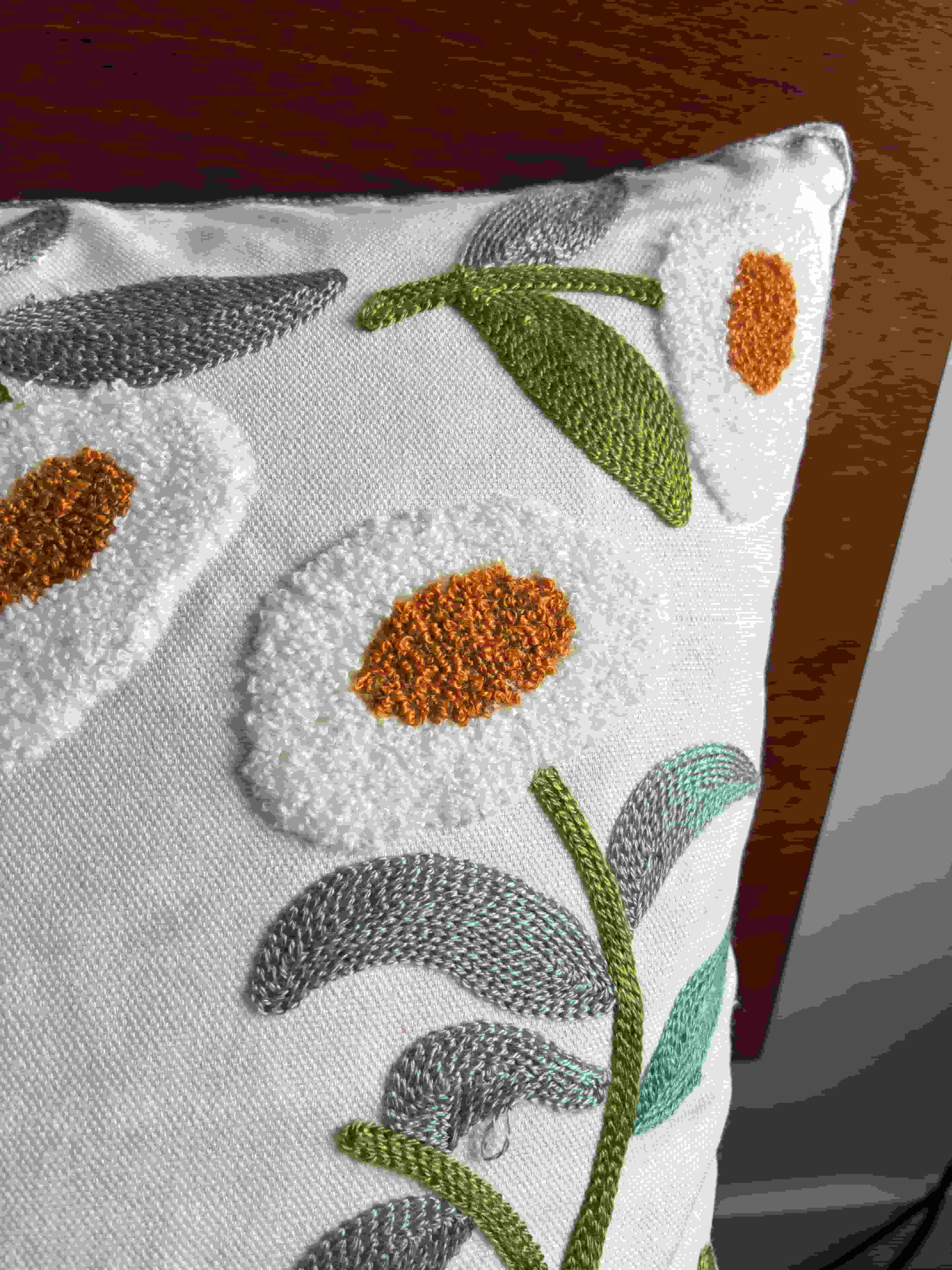 Macro shot of a white bouquet-style embroidered flower with a rust orange center, paired with stitched green and grey leaves on a white cushion cover.