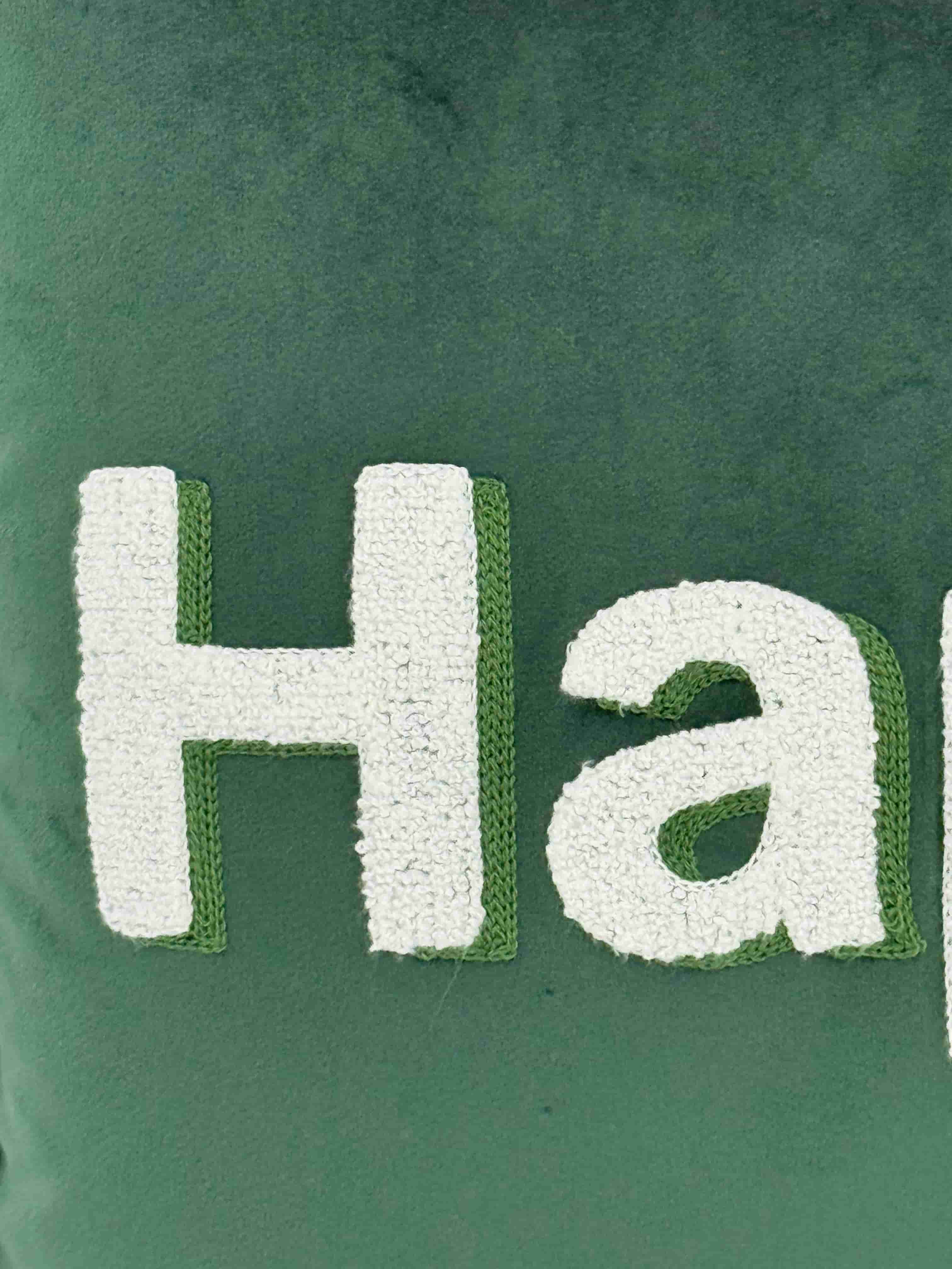 Close-up detail of white embroidered fluffy "Happy" lettering with green border stitching on an emerald green velvet cushion cover.