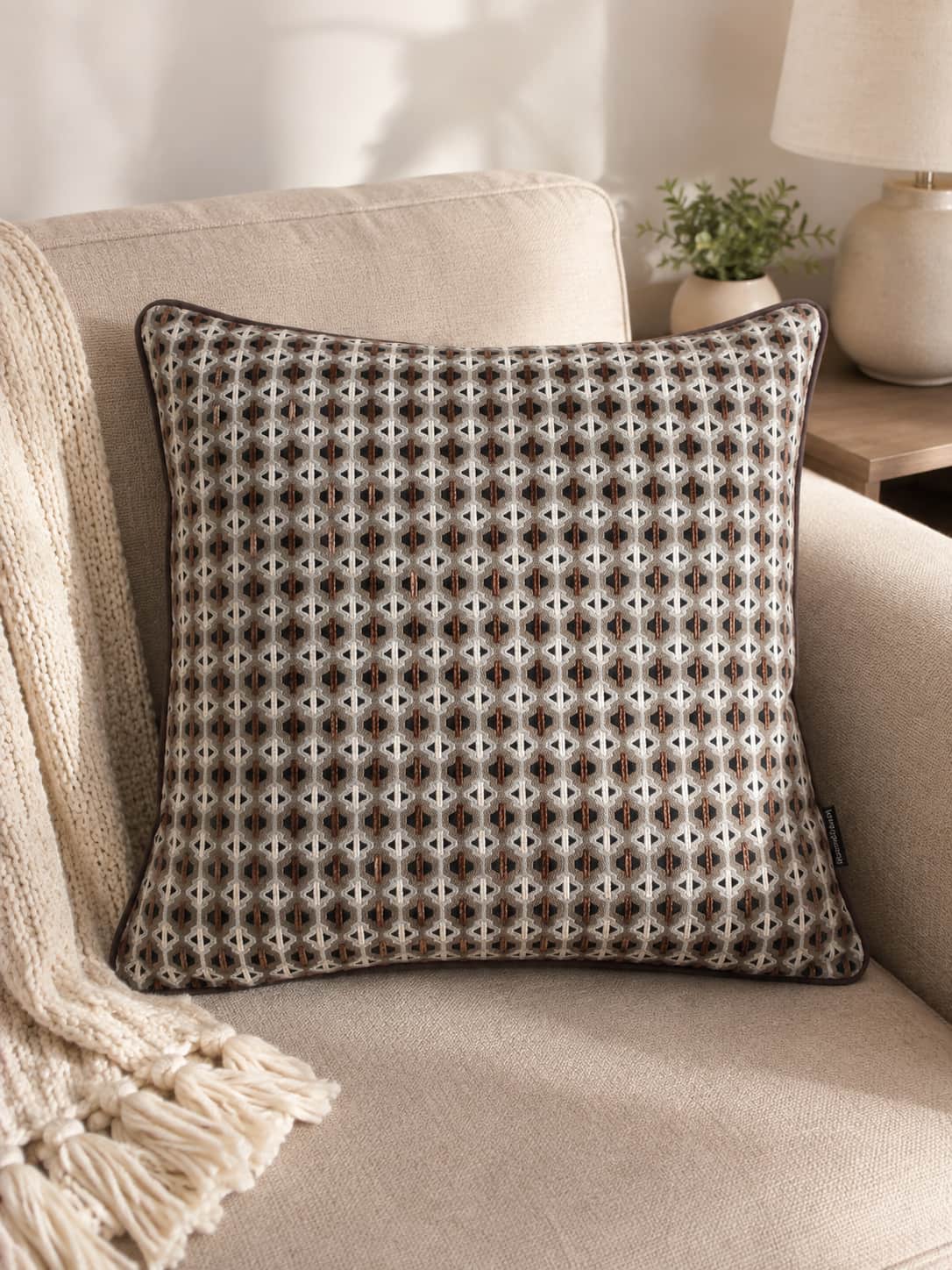 Beige and brown geometric woven throw pillow with dark piped edging, placed on a comfortable beige sofa with a cream fringed blanket and a small potted plant nearby.