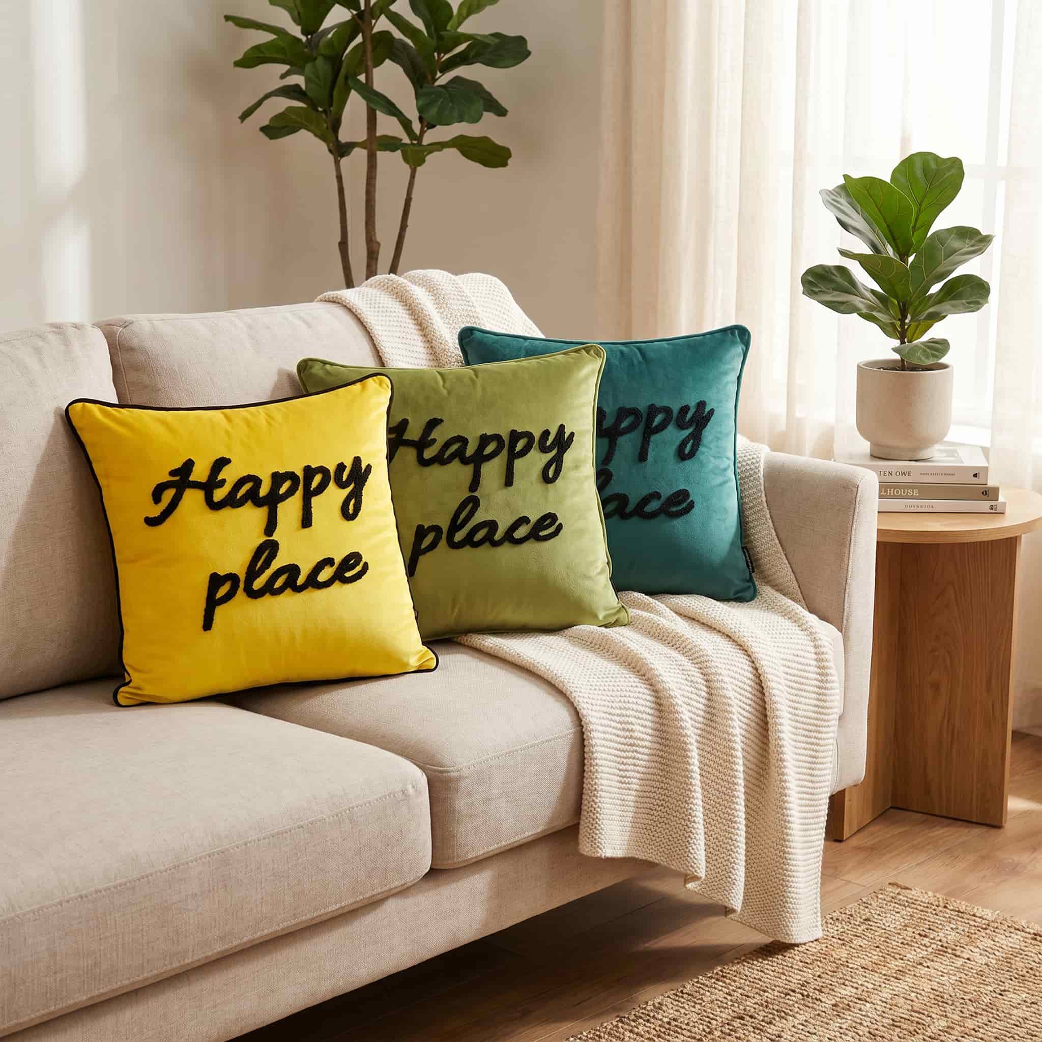 Three decorative throw pillows with 'Happy place' text in black script, displayed on a light beige sofa in a cozy living room setting. The pillows are in yellow, sage green, and teal blue, with a cream throw blanket draped over the couch.