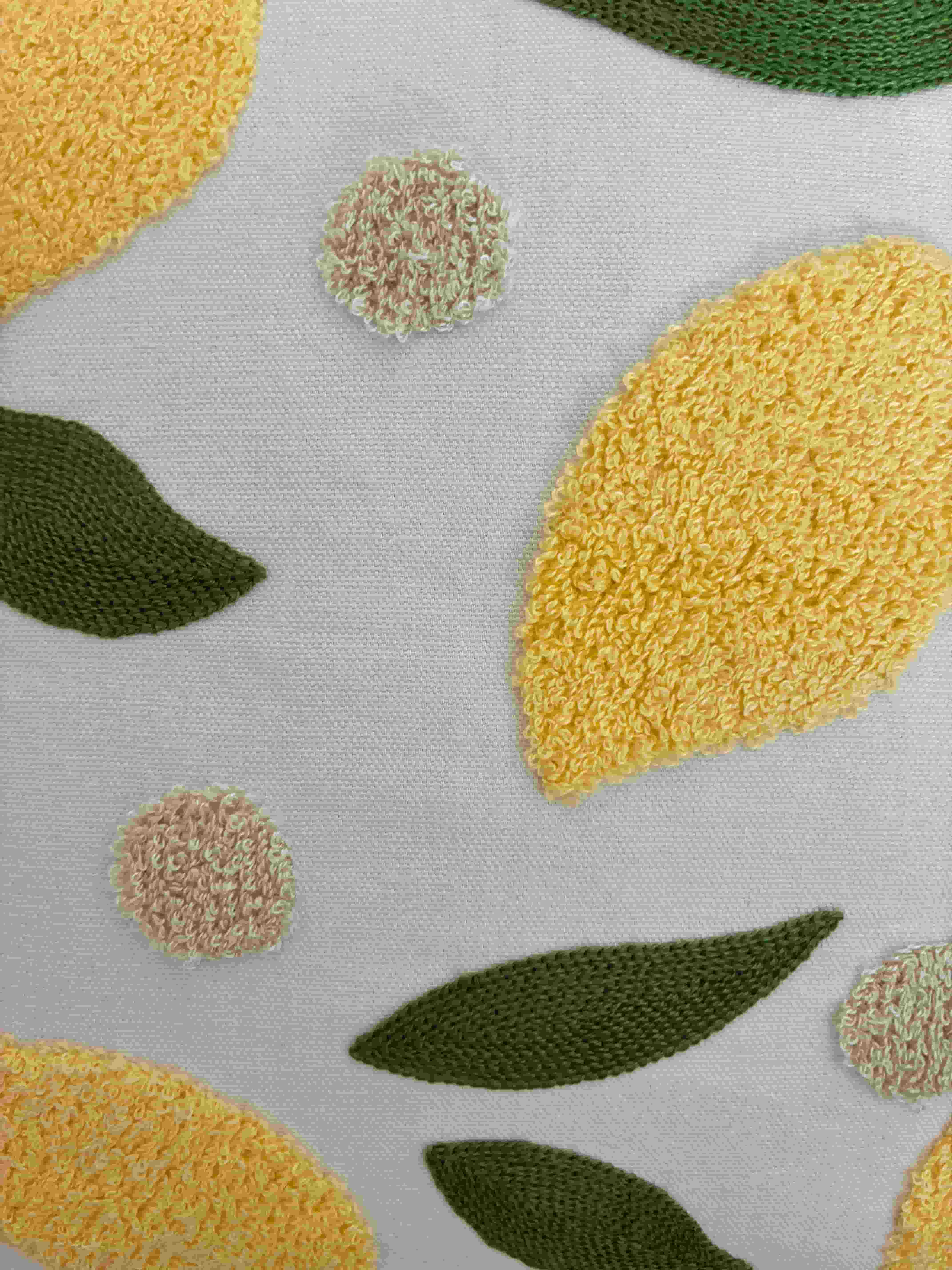 Close-up detail showing the fluffy yellow tufted lemon, green flat-stitched leaf, and cream textured pom-pom on a cream fabric cushion cover.