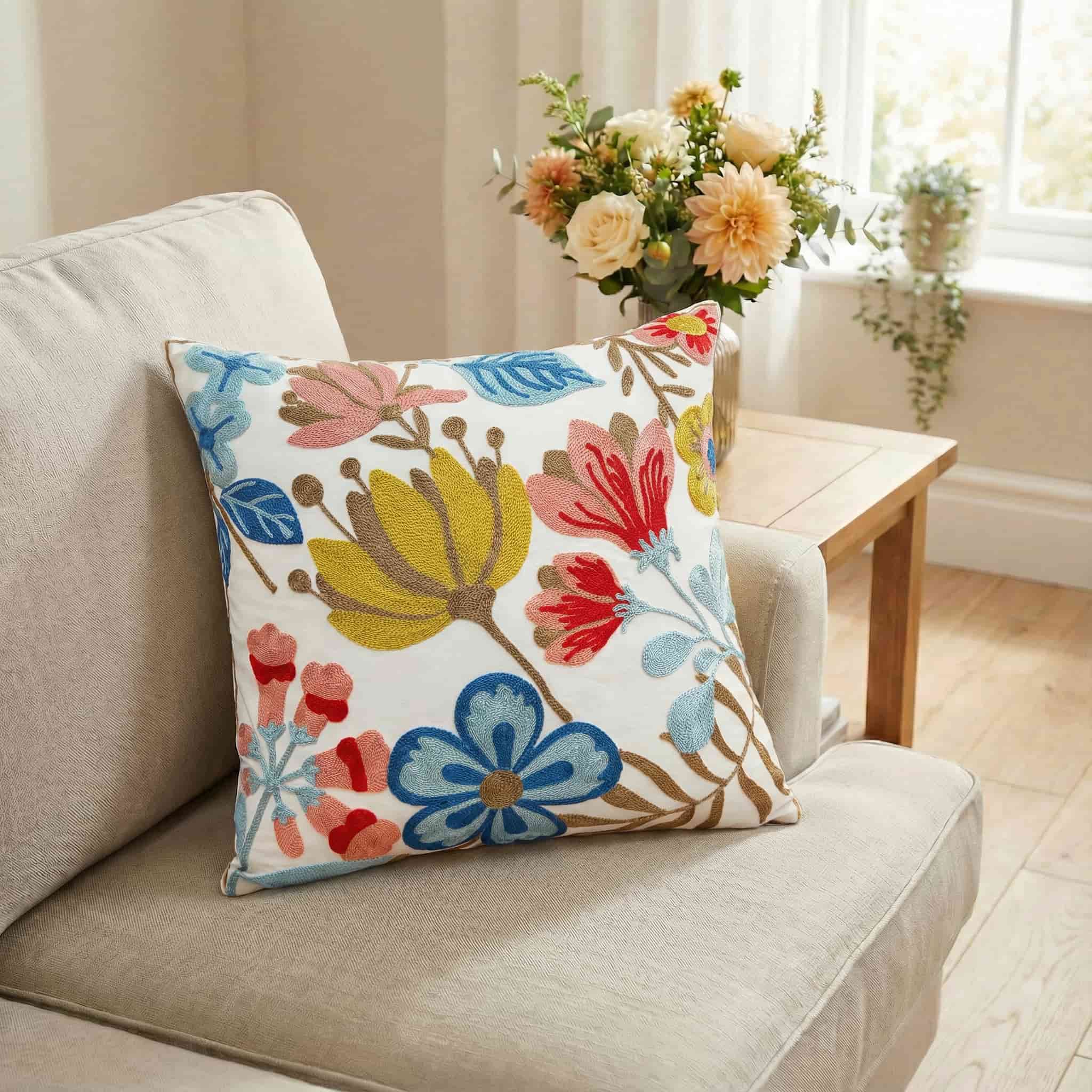 Cream throw pillow with colorful embroidered flowers in yellow, blue, pink and red, styled on a beige armchair next to a table with a large vase of fresh cut flowers.