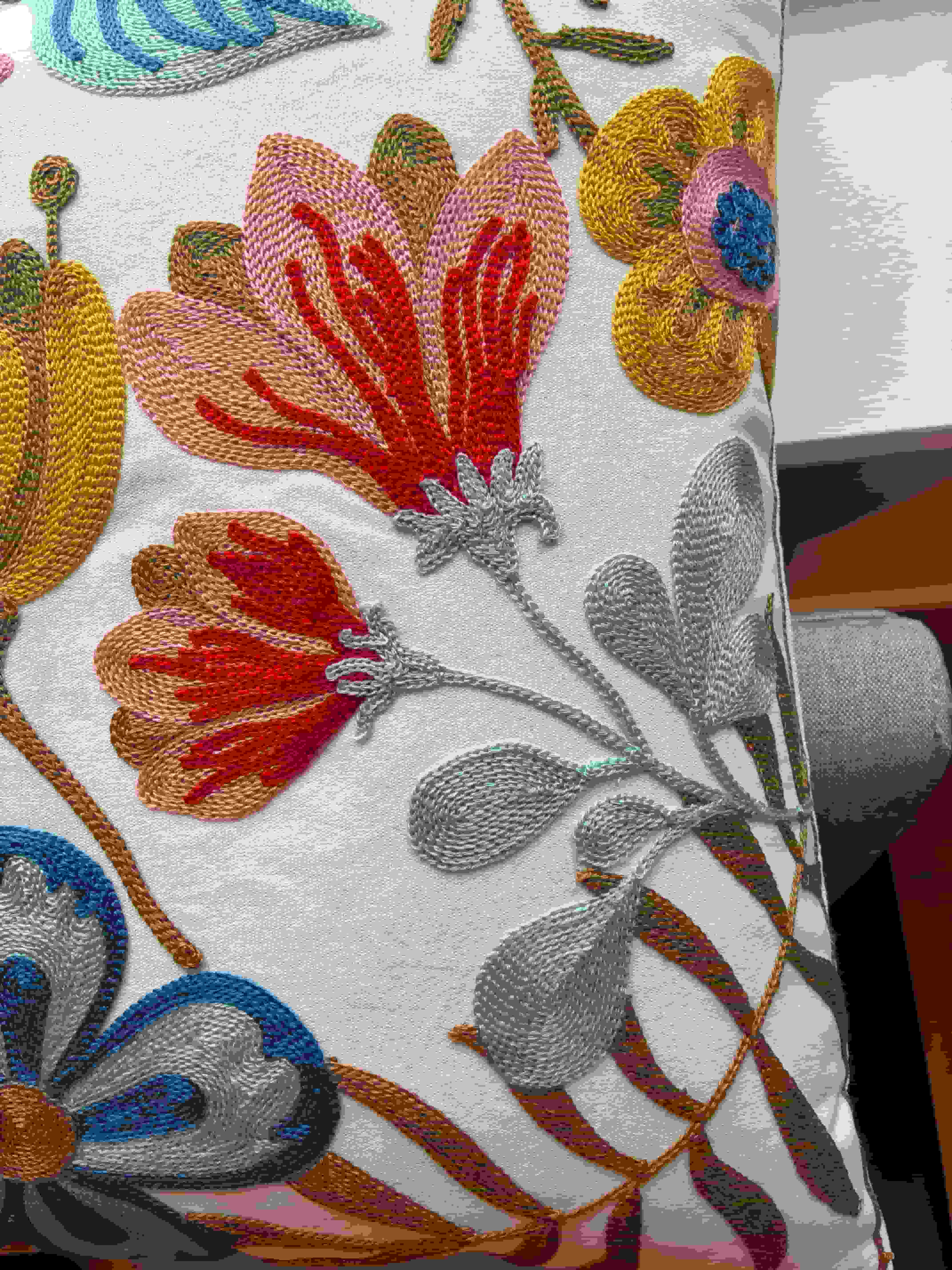 Close-up macro shot of colorful embroidered flowers on a cream cushion cover, showcasing detailed stitching in mustard yellow, vibrant red, and light blue.