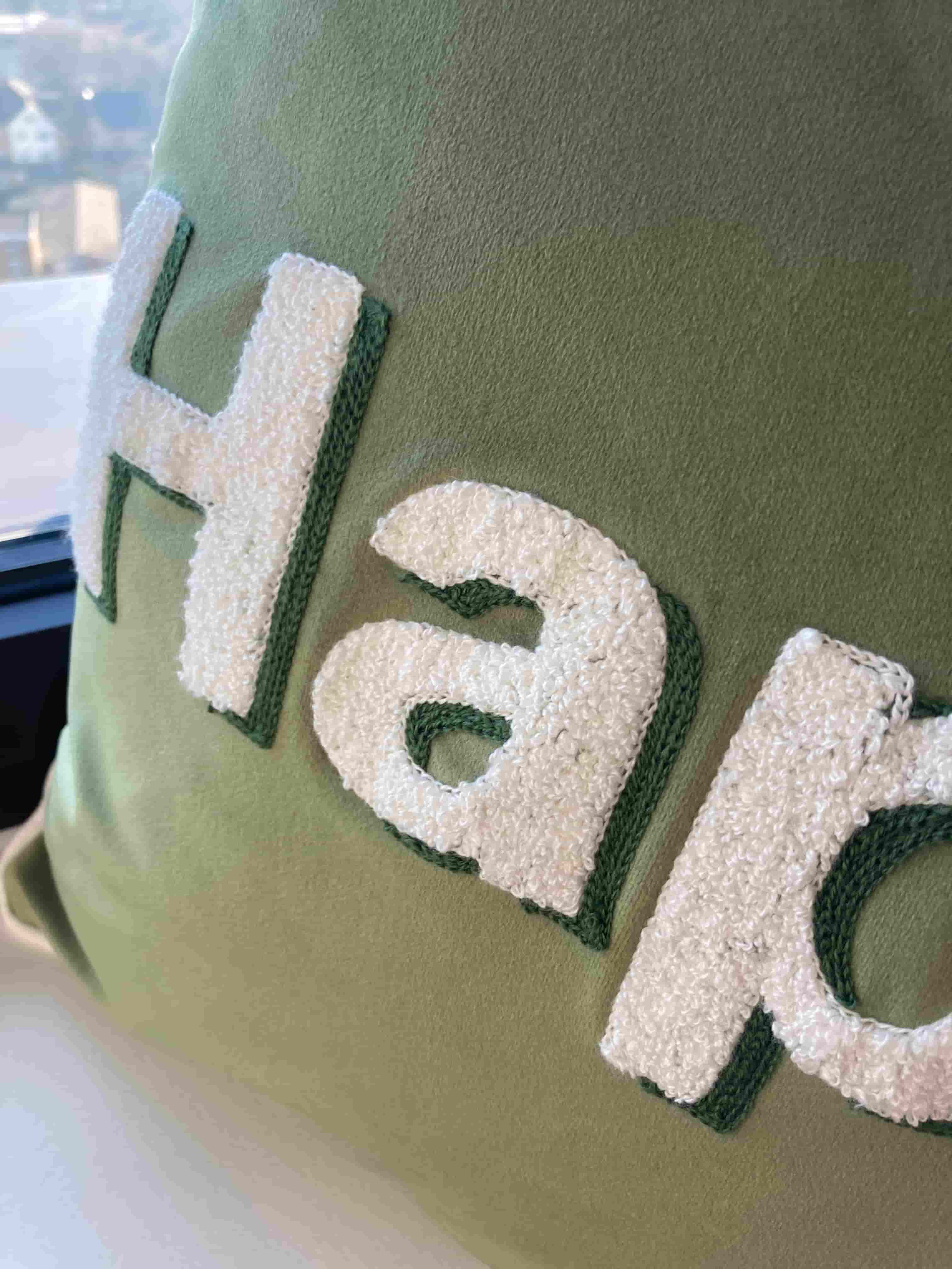 Close-up detail of white boucle-style embroidered "Happy" text with green border stitching on an olive green velvet cushion cover.