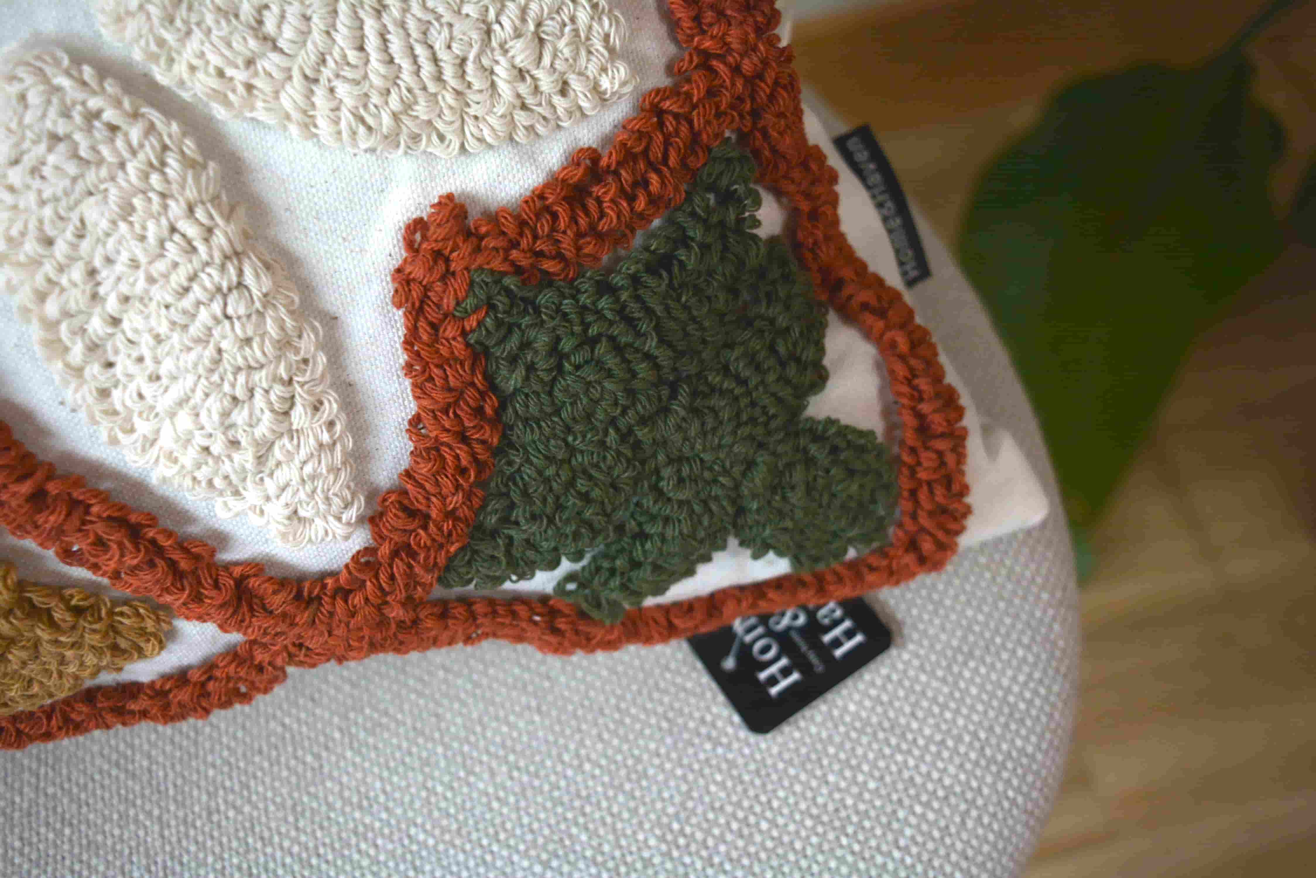 Macro shot showing a dark olive green tufted leaf pattern and thick rust-colored loop yarn border on a white cushion cover with a black brand label.