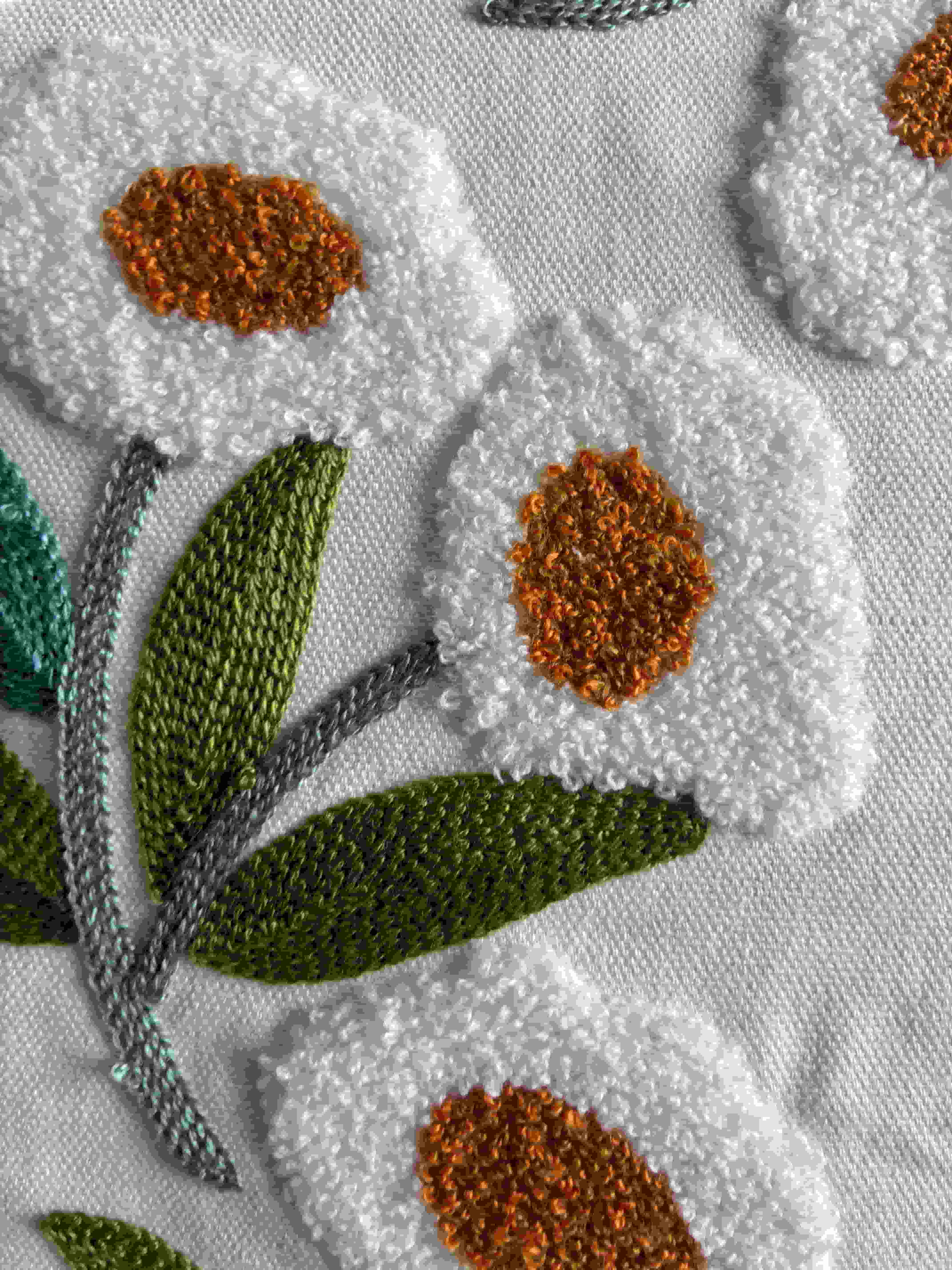 Close-up of textured white embroidered flower petals with raised orange centers and stitched sage green leaves on a cream fabric cushion cover.