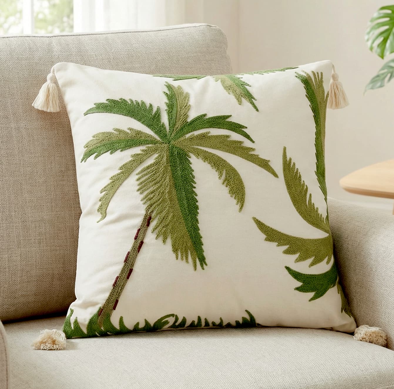 Cream throw pillow with green embroidered palm tree design and four cream corner tassels, placed on a beige sofa in a bright room with a green plant in the background.
