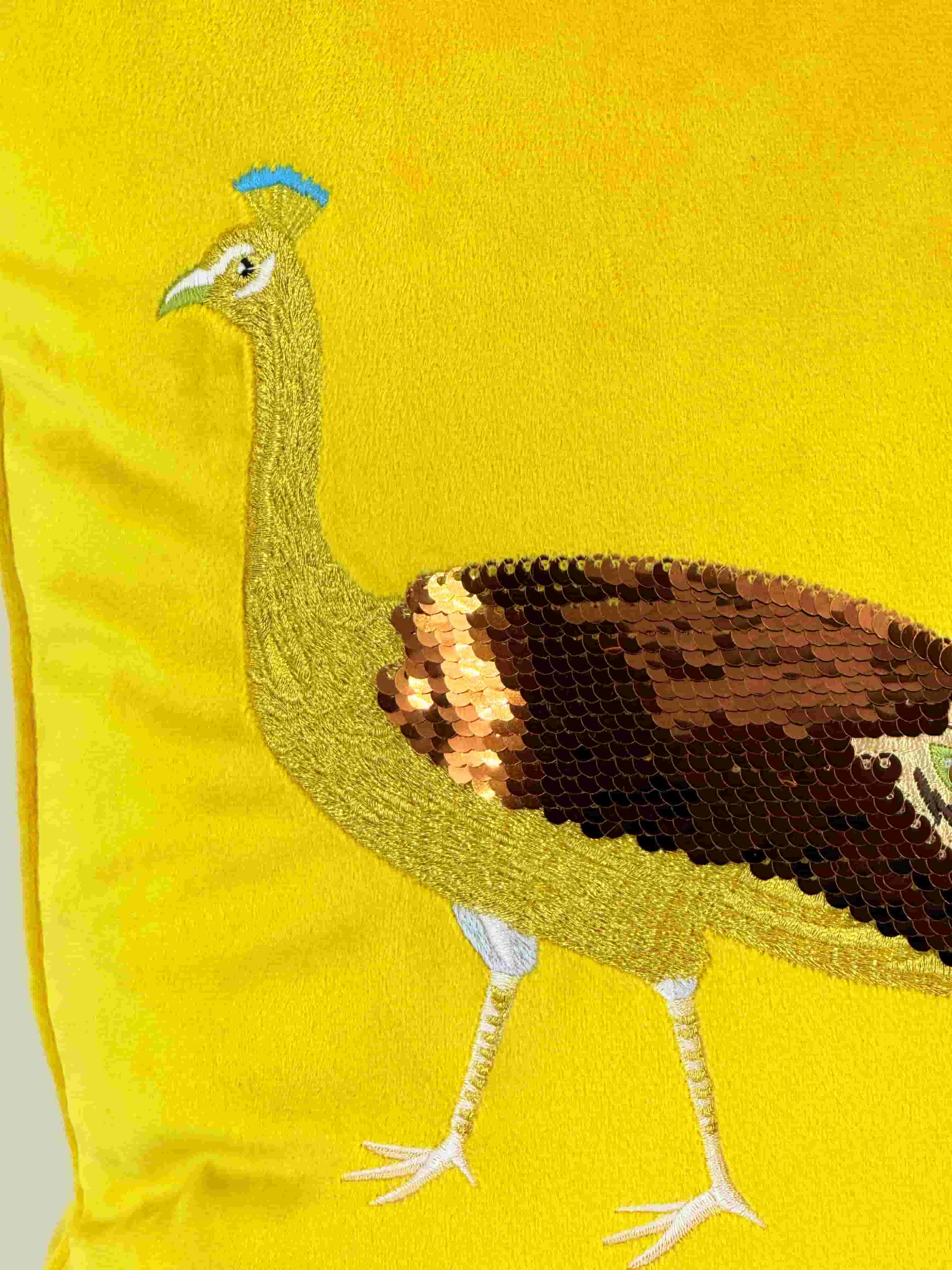 Close-up detail of a gold embroidered peacock head and neck, showing dark brown and copper sequin detailing on the wing, set against a yellow velvet cushion cover.