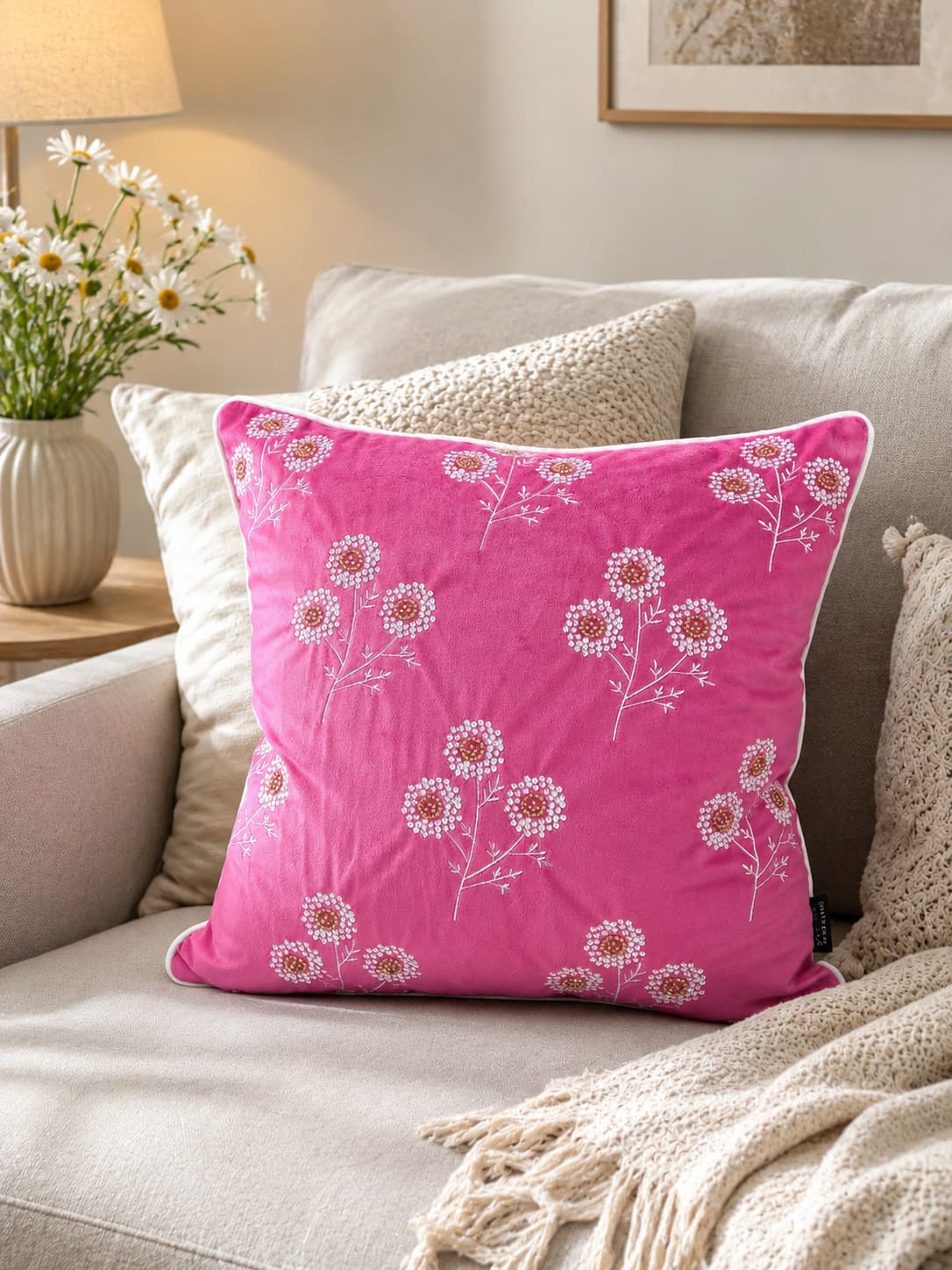 Bright pink velvet throw pillow with white embroidered flowers and gold bead centers, styled on a beige sofa with a white fringe blanket and a vase of fresh daisies.