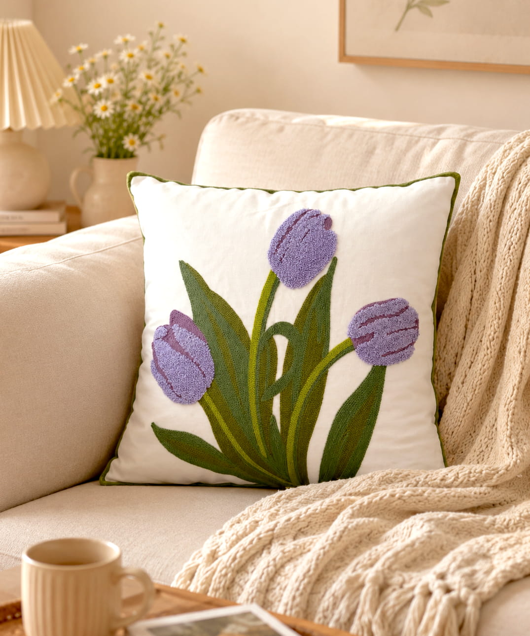 Cream throw pillow with three plush lavender purple tufted tulips and green embroidered leaves, styled on a beige sofa in a bright living room.