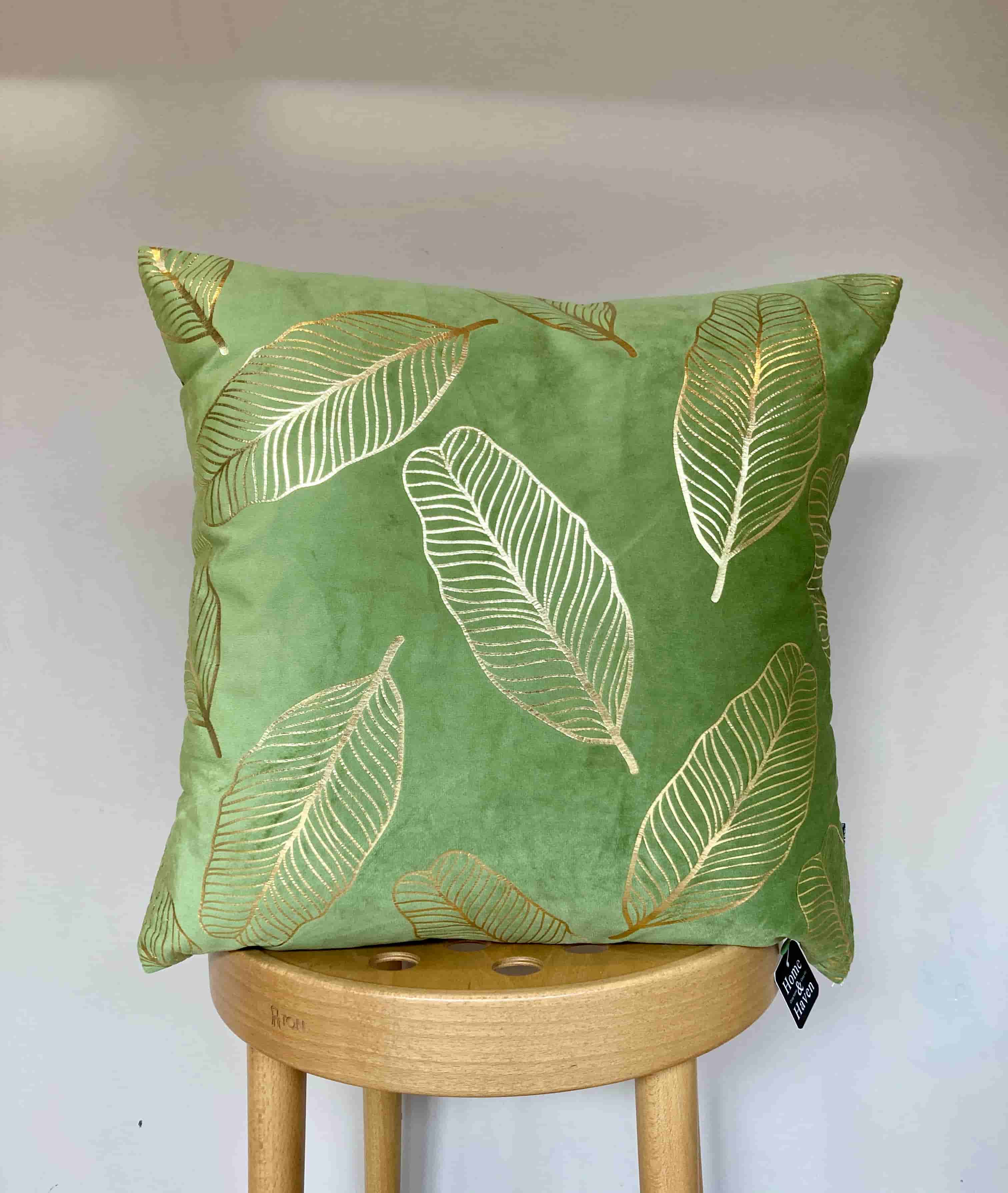 Sage green velvet throw pillow with elegant gold print, displayed upright on a light wood stool against a white background.