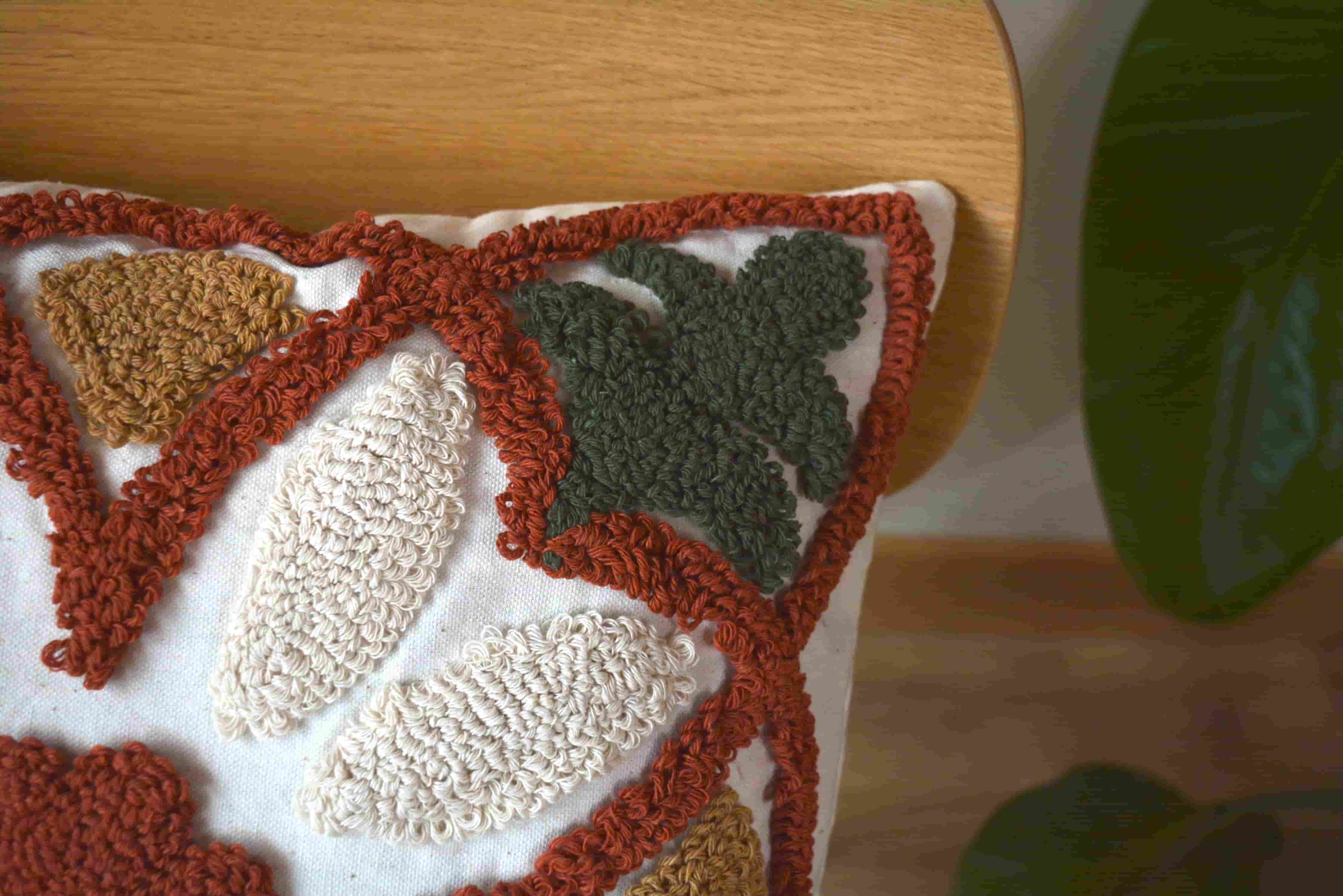 Detailed view of mustard yellow and dark green tufted leaves, cream petals, and thick rust-colored loop yarn borders on a cream pillow.