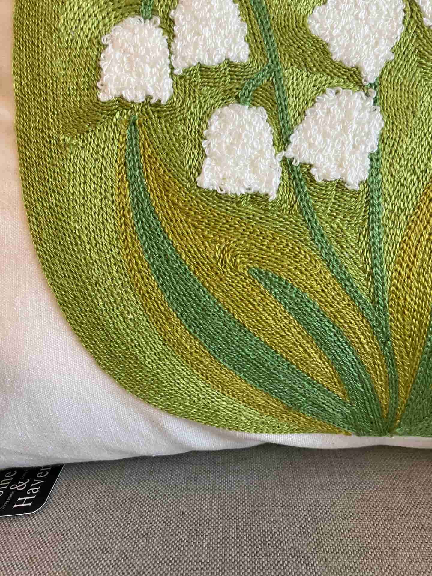 Close-up shot of white tufted lily of the valley flowers and green stitched leaves on a cream fabric cushion cover, with a black Home & Haven logo tag visible at the bottom edge.