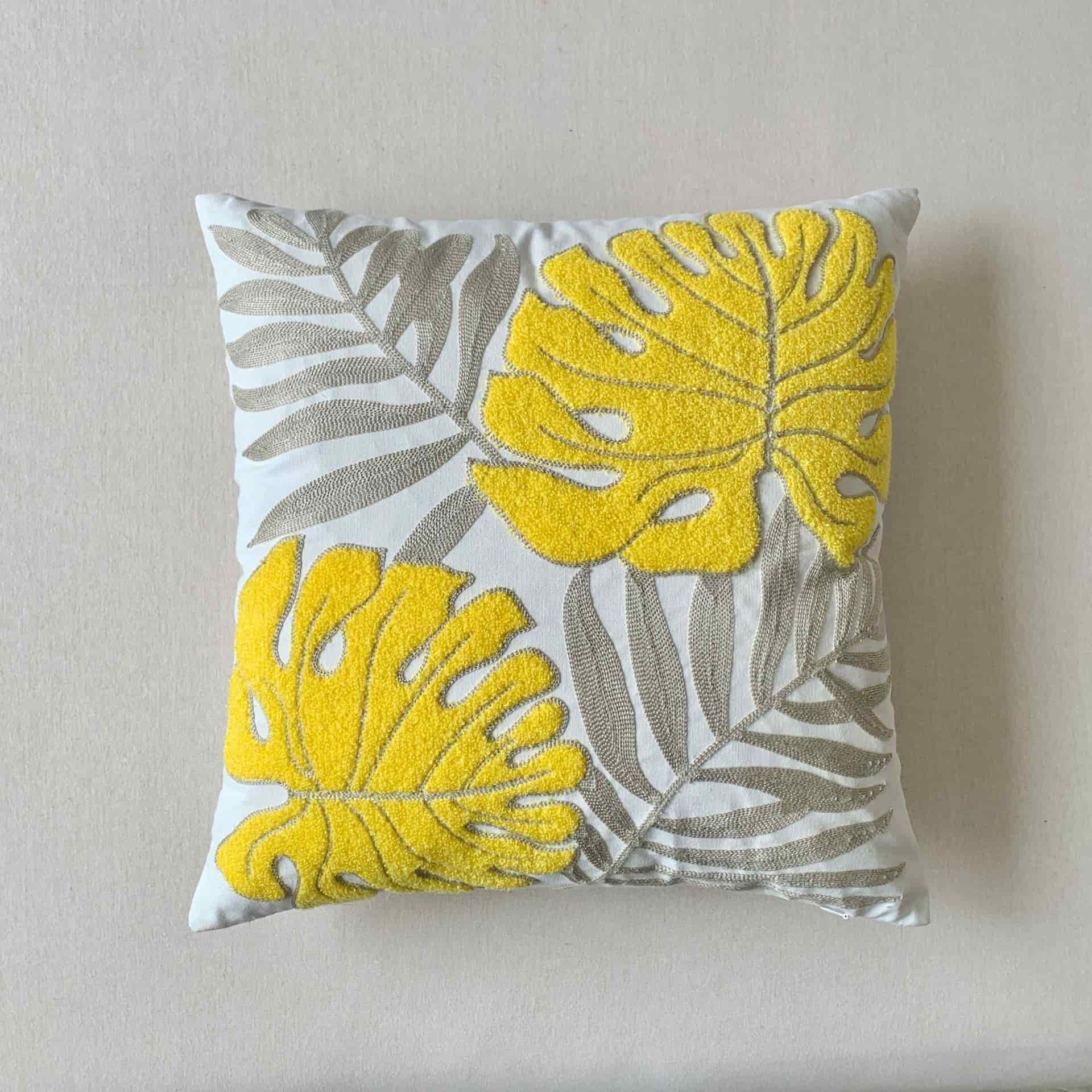 Square off-white cushion cover featuring textured yellow embroidery of monstera leaves and flat-stitched grey fern leaves, placed on a clean white surface.
