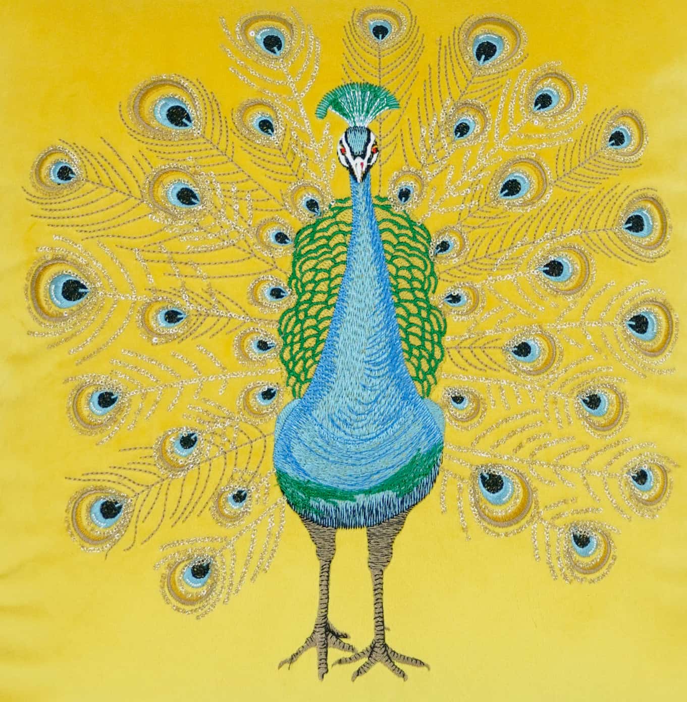 Flat lay of a bright yellow cushion cover featuring exquisite embroidery of a blue and green peacock with a fully spread tail and metallic thread details.