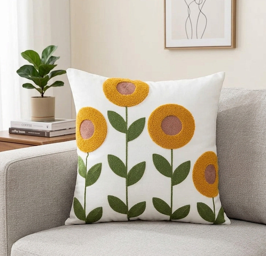 Minimalist square cushion cover with fluffy yellow embroidered flowers and green stems, placed on a textured grey sofa next to a potted plant.
